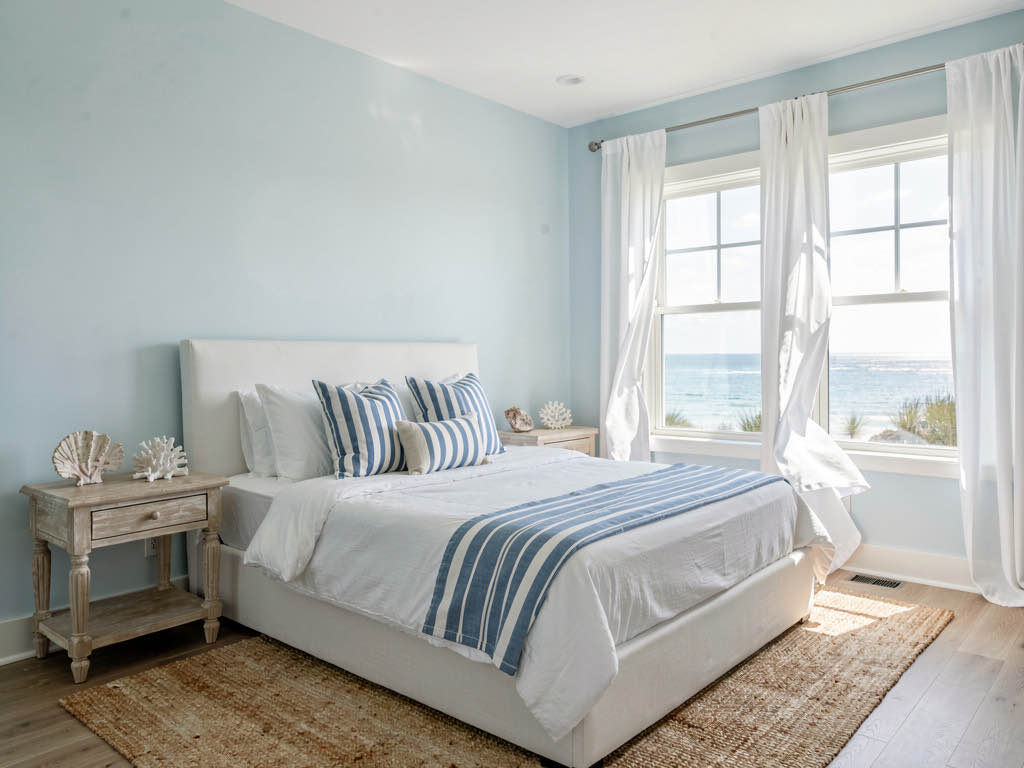 A coastal bedroom with a white shiplap wall, a cane headboard, light blue linen bedding, and a jute rug under the bed